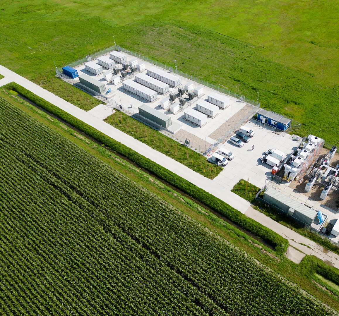 Aerial view of a fenced battery energy storage system site with rows of white battery containers, inverter skids and a grid substation yard